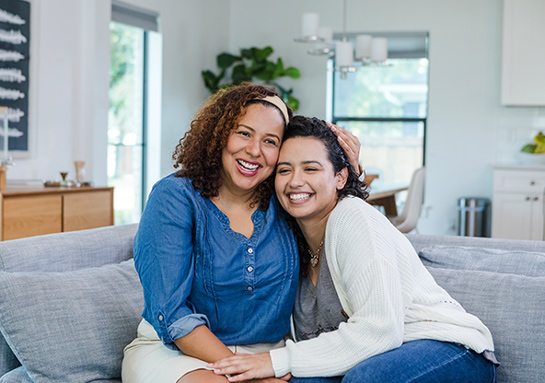 A woman and her adult daughter are hugging on the couch